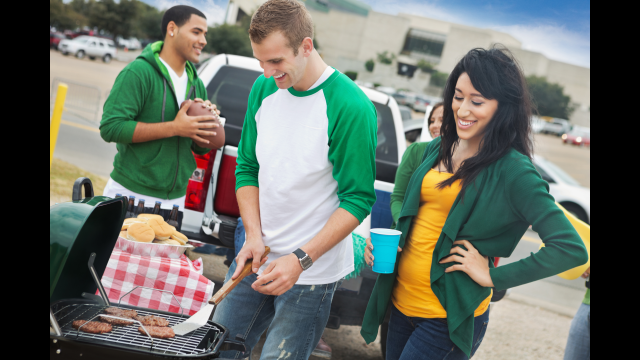 people smiling and looking down at cooking grill in front person with football in a parking lot